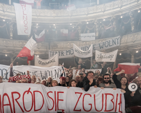 Tłum protestujących w przestrzeni widowni dużej sceny teatru. Trzymają biało-czerwone flagi oraz transparenty z hasłami i duży baner z napisem „Naród się zgubił”. Część osób unosi zaciśnięte pięści, jedna trzyma megafon. Tło jest lekko zadymione.